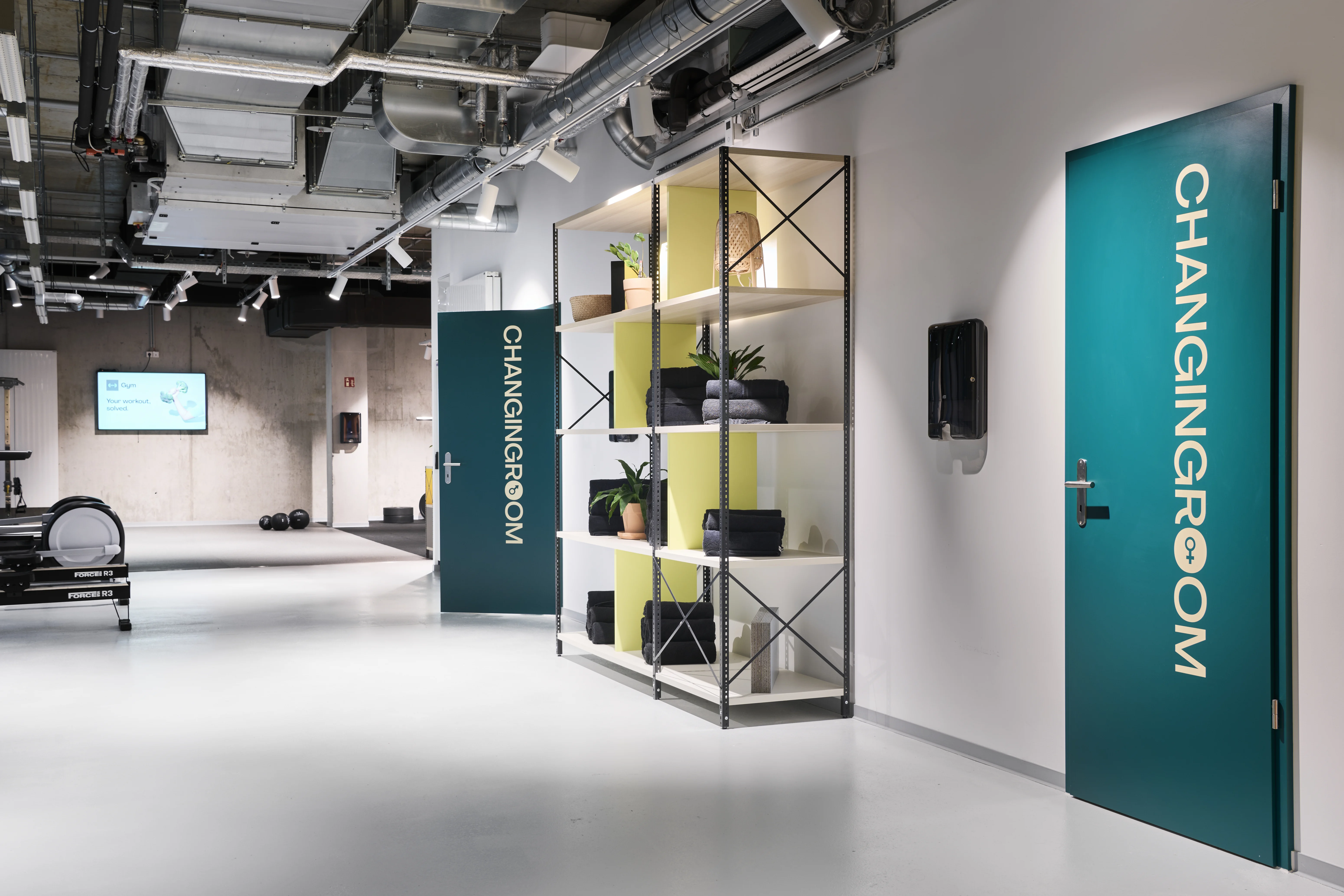 Entrance zone of the HELLOFRESH GYM with petrol-green doors labelled Changingroom, an open metal shelf with towels and plants and the training area in the background.