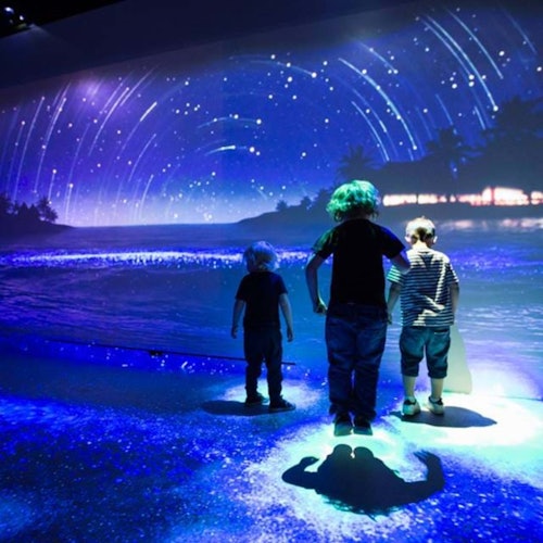 Three children stand in front of a large, immersive projection of a nighttime beach scene with stars, waves, and silhouettes of trees.