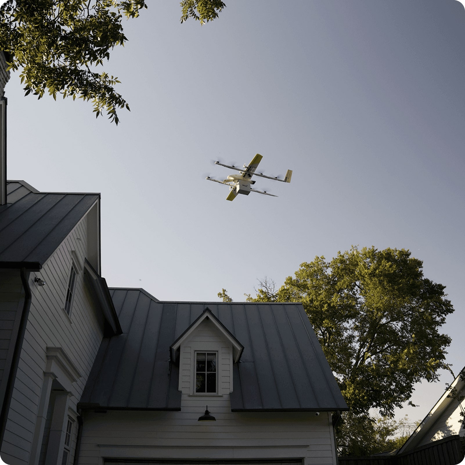 A drone with multiple propellers flies over a suburban neighborhood, casting a shadow on a house with a gray metal roof and surrounded by lush green trees under a clear blue sky.