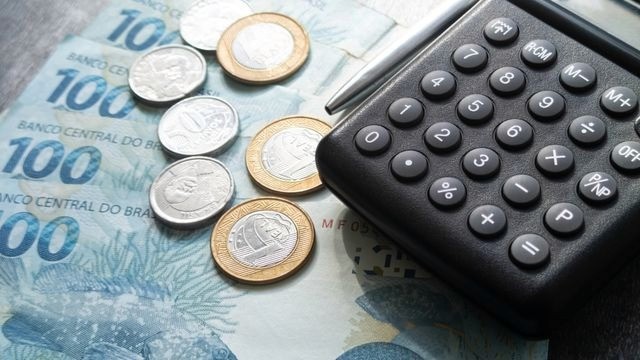 Notas e moedas de real sob uma caneta e uma calculadora.