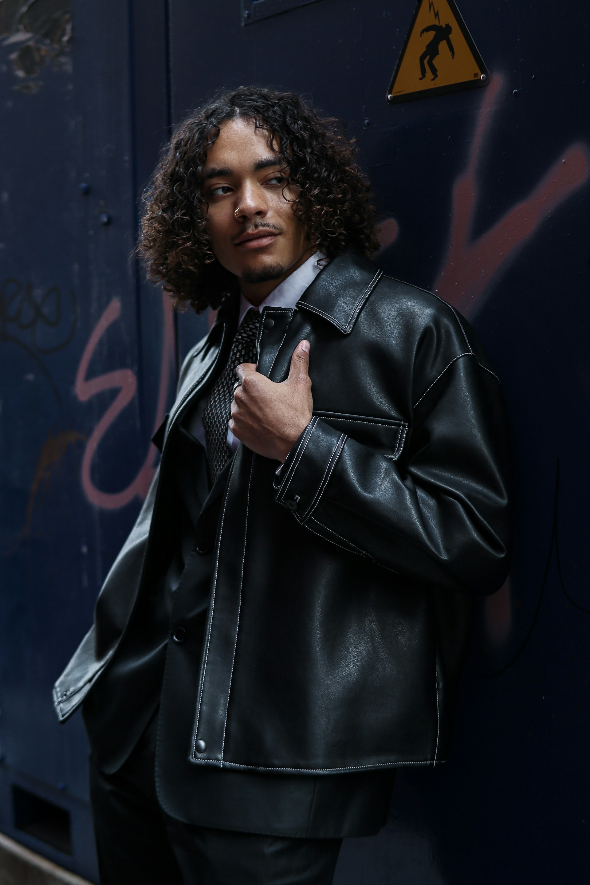 A man with curly hair and a nose ring stands against a dark blue wall with graffiti. He is wearing a white dress shirt, a patterned tie, and a black leather jacket with white contrast stitching.