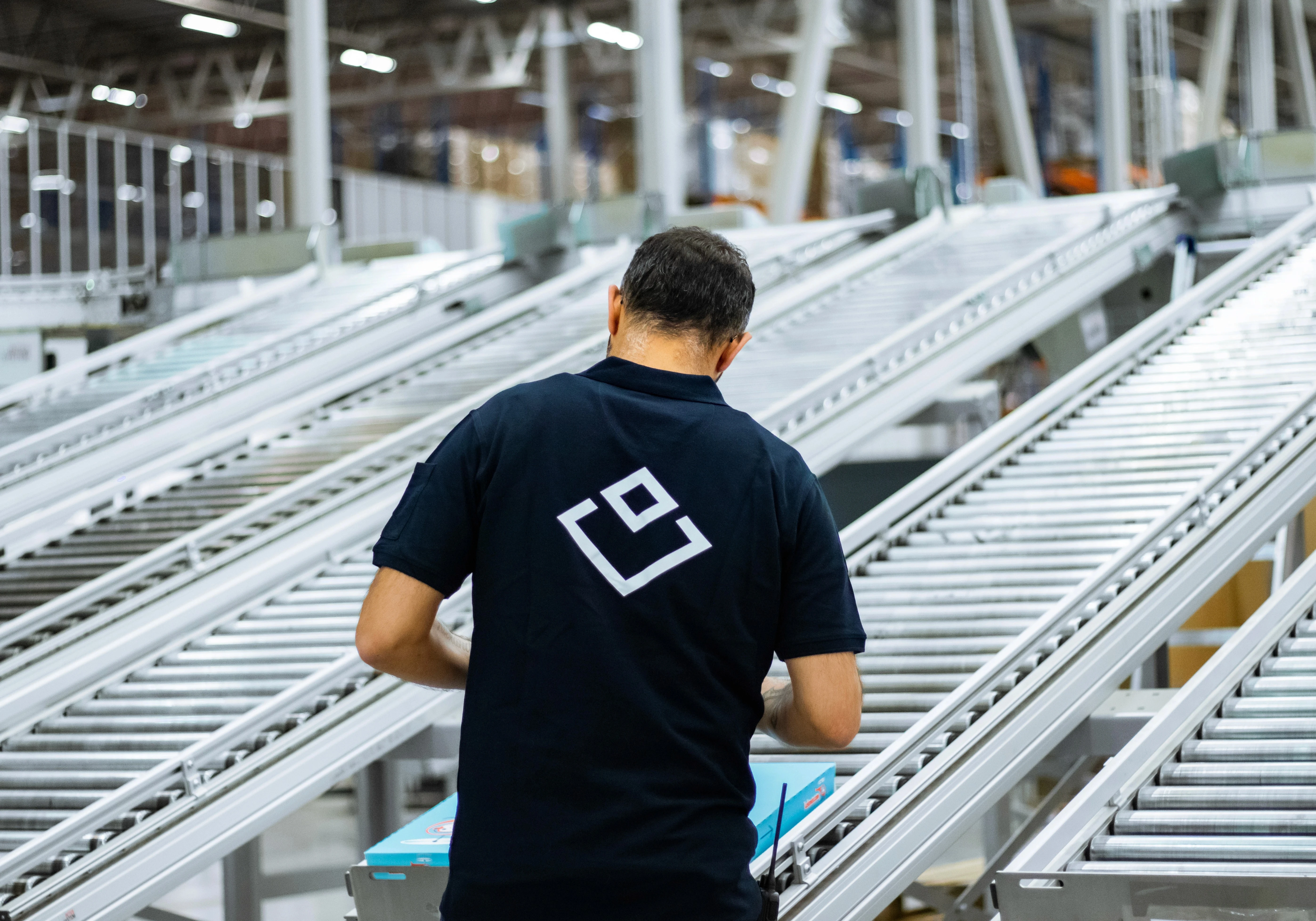 En man i svart skjorta med logotyp arbetar vid transportband på en fabrik, nära bild.