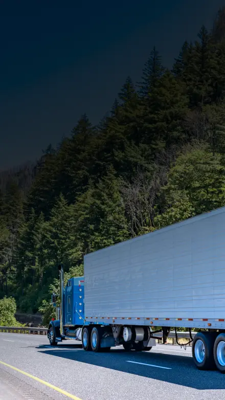 Blue semi-truck with a white trailer driving through a scenic mountain highway surrounded by pine trees, representing successful trucking operations insured by GIA Group, LLC.