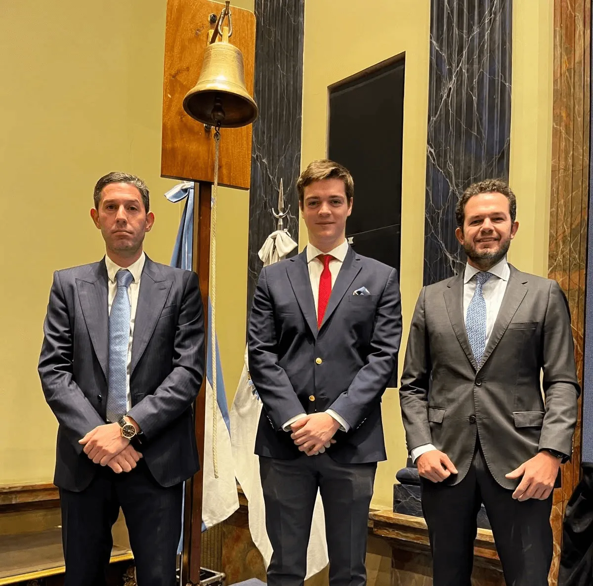 Tres personas de traje frente a una gran campana montada, con columnas de mármol al fondo.