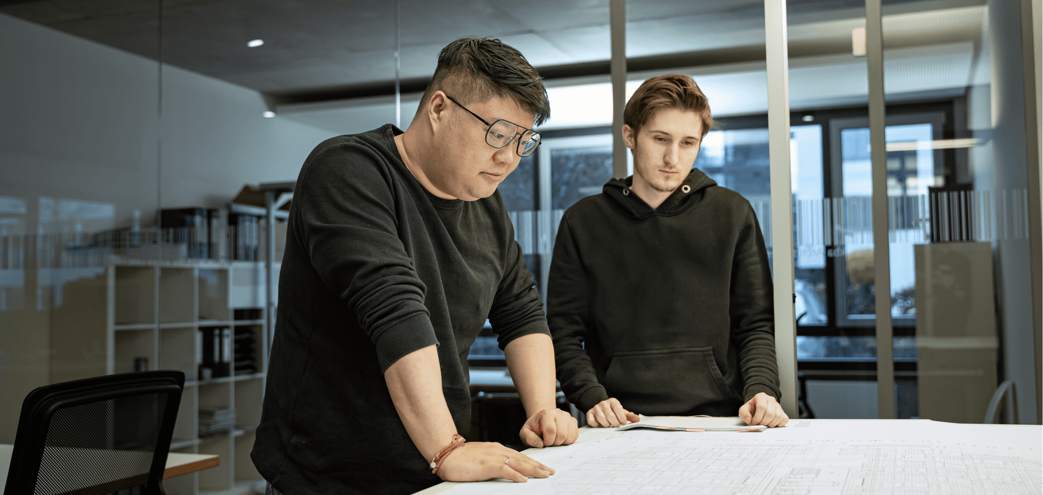 Zwei Mitarbeitende der kba Architekten und Ingenieure GmbH stehen gemeinsam an einem großen Tisch und prüfen detaillierte Baupläne; der Raum ist modern eingerichtet mit Glaswänden und Regalen im Hintergrund.