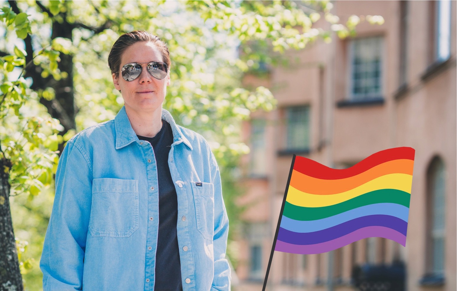 Tuulia Virhiä with short hair wearing sunglasses and a light blue shirt standing outdoors with a rainbow pride flag