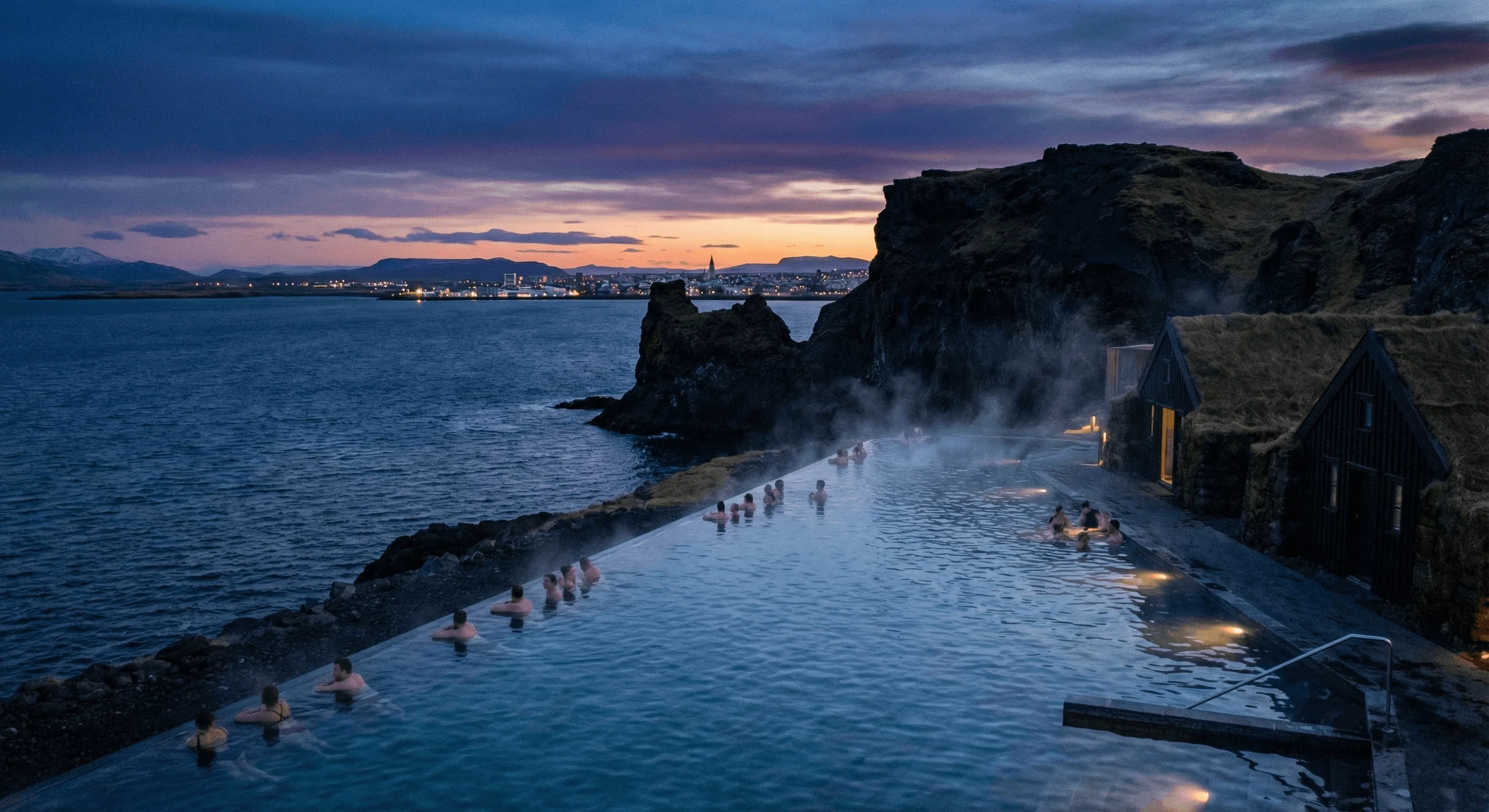 A geothermal infinity pool overlooking the ocean at twilight with people soaking in the warm water.