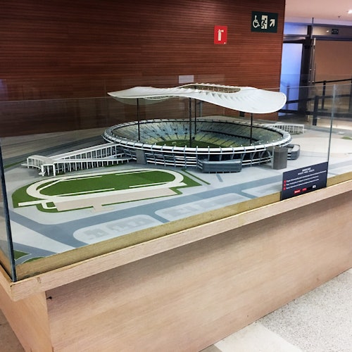 Un model detaliat la scară al unui stadion cu locuri de ședere, o pistă și un acoperiș unic ondulat, închis într-o vitrină de sticlă în interior.