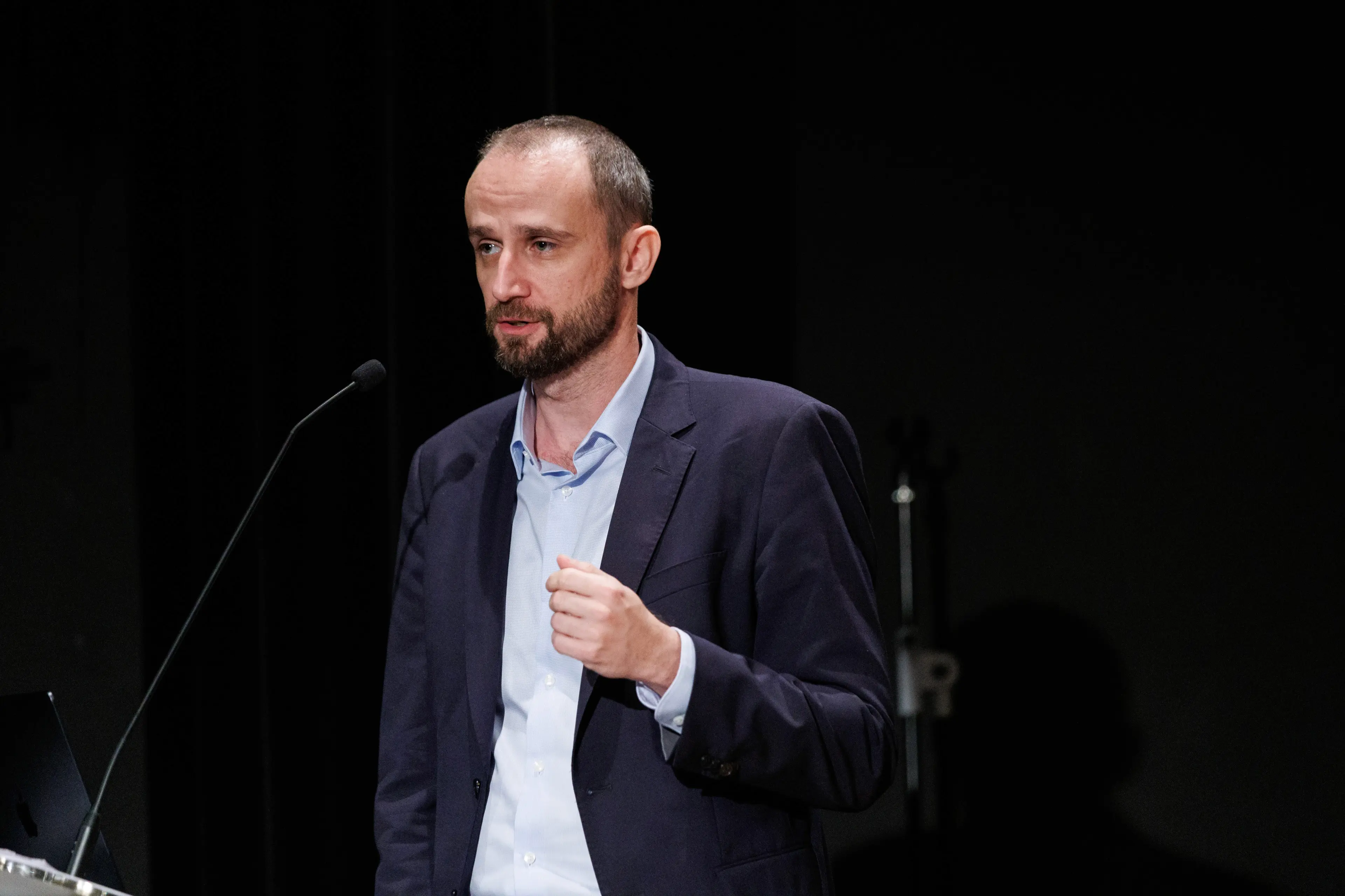 A man in a suit stands on stage, speaking into a microphone with a thoughtful expression.