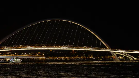 A large, modern arched bridge in China illuminated with golden lights at night, reflecting over a dark body of water with a city skyline in the distance.