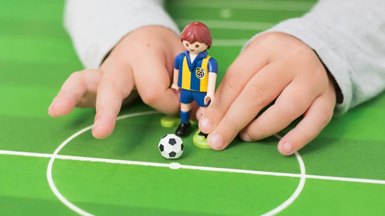 Close-up of children's hands playing with a Playmobil football figure on a miniature pitch. The scene symbolises the playful and child-friendly concept of the funino festival league in kids' football.