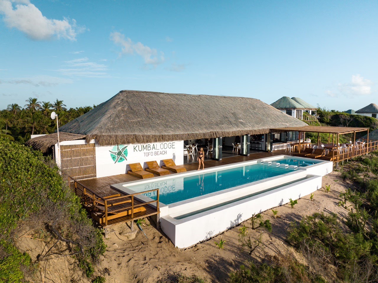 Vista elevada de una propiedad frente a la playa con una piscina infinita, pérgola de madera y dunas que conducen al océano.