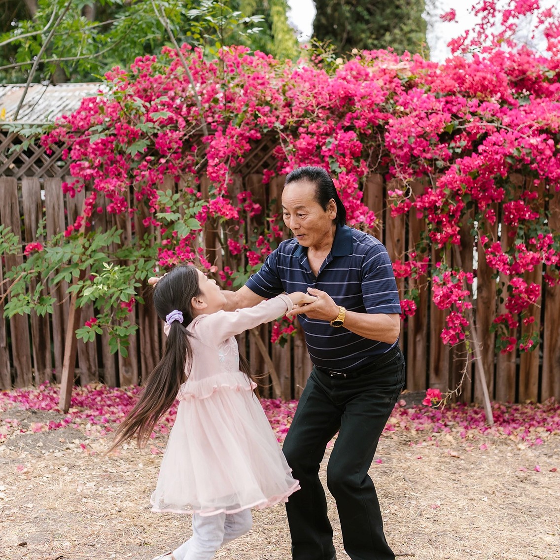 An elderly man dancing with his young granddaughter in front of some vibrant pink flowers