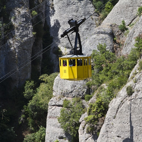 在山峦起伏的景观中，一辆黄色缆车悬挂在岩石峭壁和绿树之间。