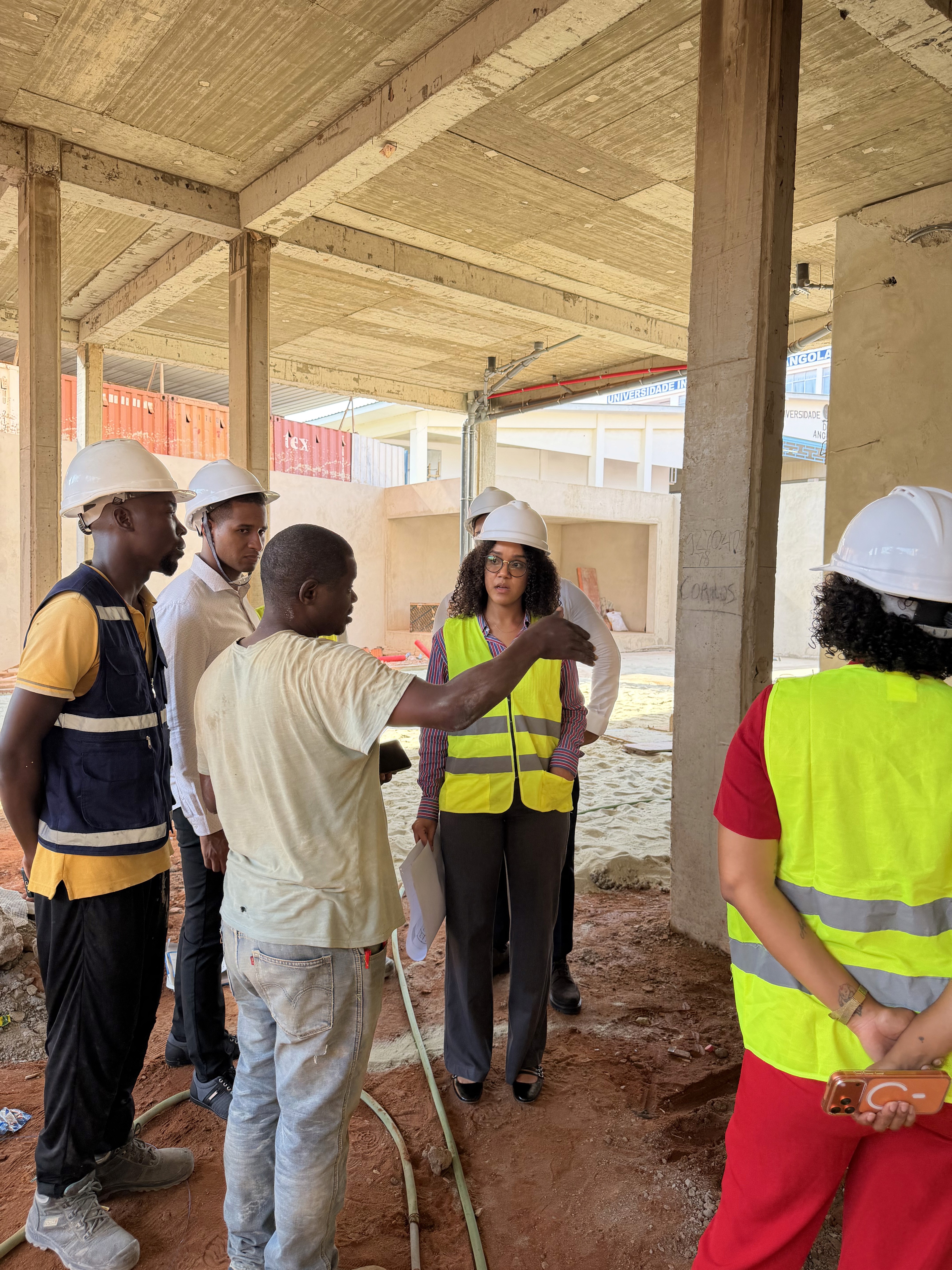 Modern office with three professionals in discussion, surrounded by computers and architectural drawings. Glass walls and pendant lights create an open, collaborative atmosphere.