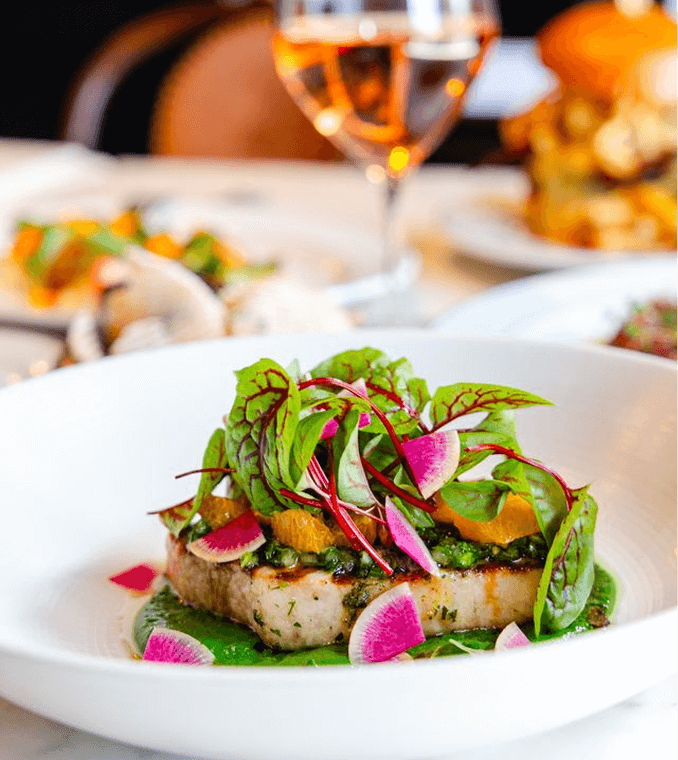 A plated dish featuring a seared fish fillet topped with microgreens, sliced radishes, and orange segments, with a green sauce, served on a white plate. A glass of rosé in the background.