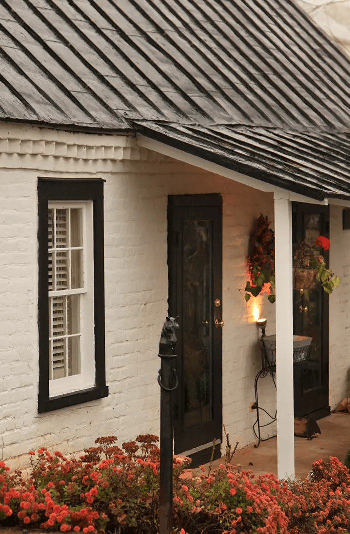 A charming, rustic white-brick cottage at The Estate at Meander features a black metal roof, dark-framed windows, and a black door, accented by vibrant red flowers and warm, ambient lighting.