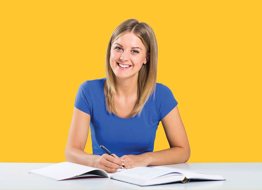 A woman in a blue shirt sits at a desk taking notes from a text book
