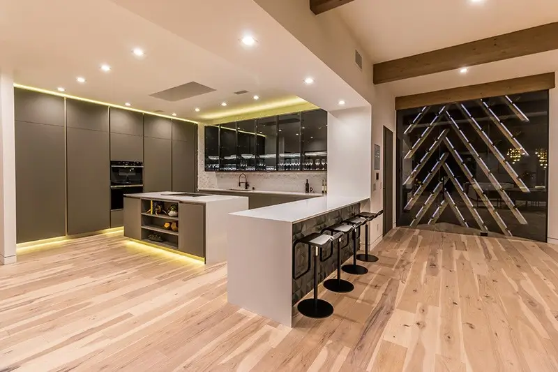 Contemporary kitchen installation with sleek gray cabinets, a stylish island, and a unique illuminated wine storage wall on wooden flooring