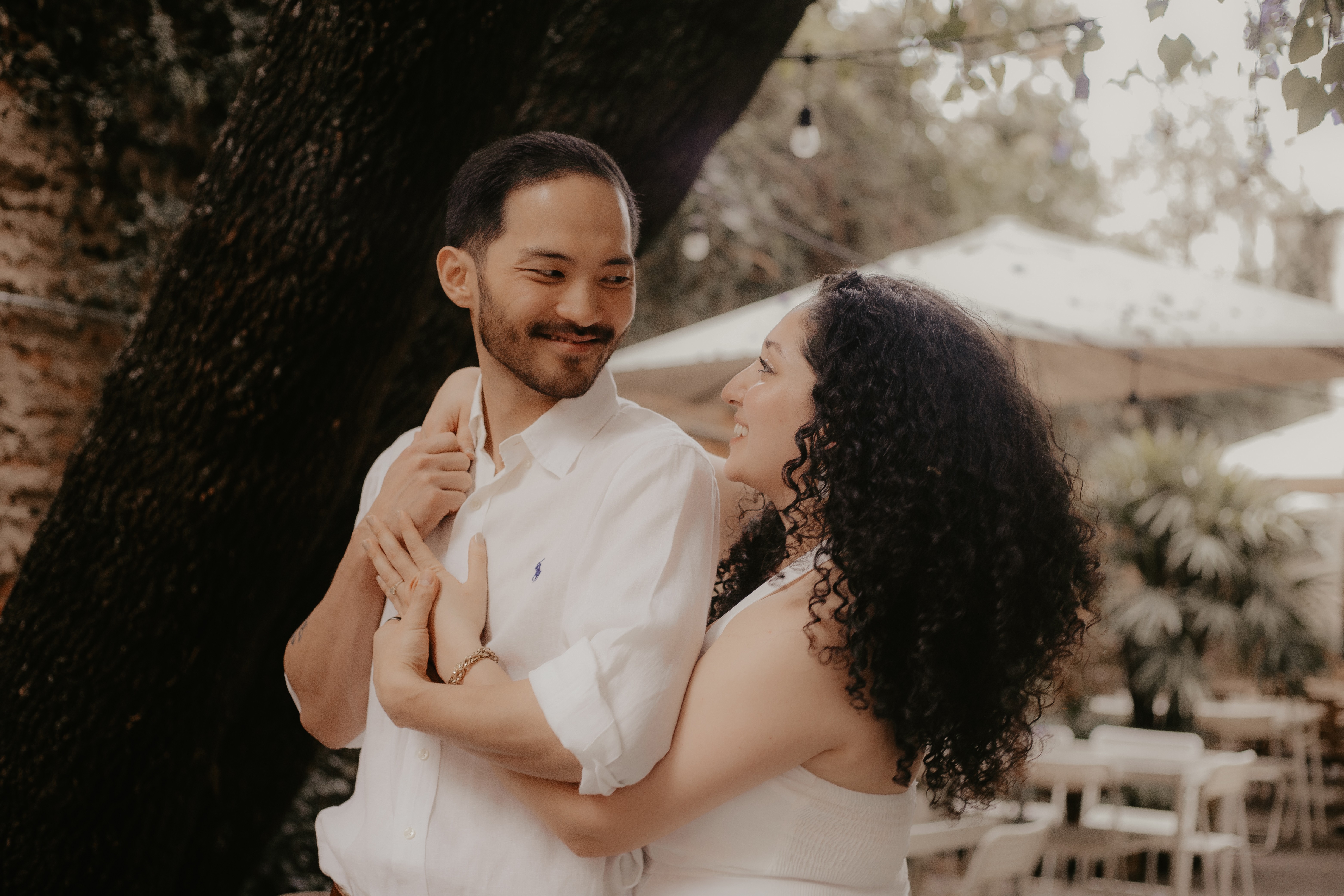 A couple holds hands, standing on a rocky beach under a large cave arch, gazing at the tranquil ocean during sunset. The sky is softly lit in pastel hues.