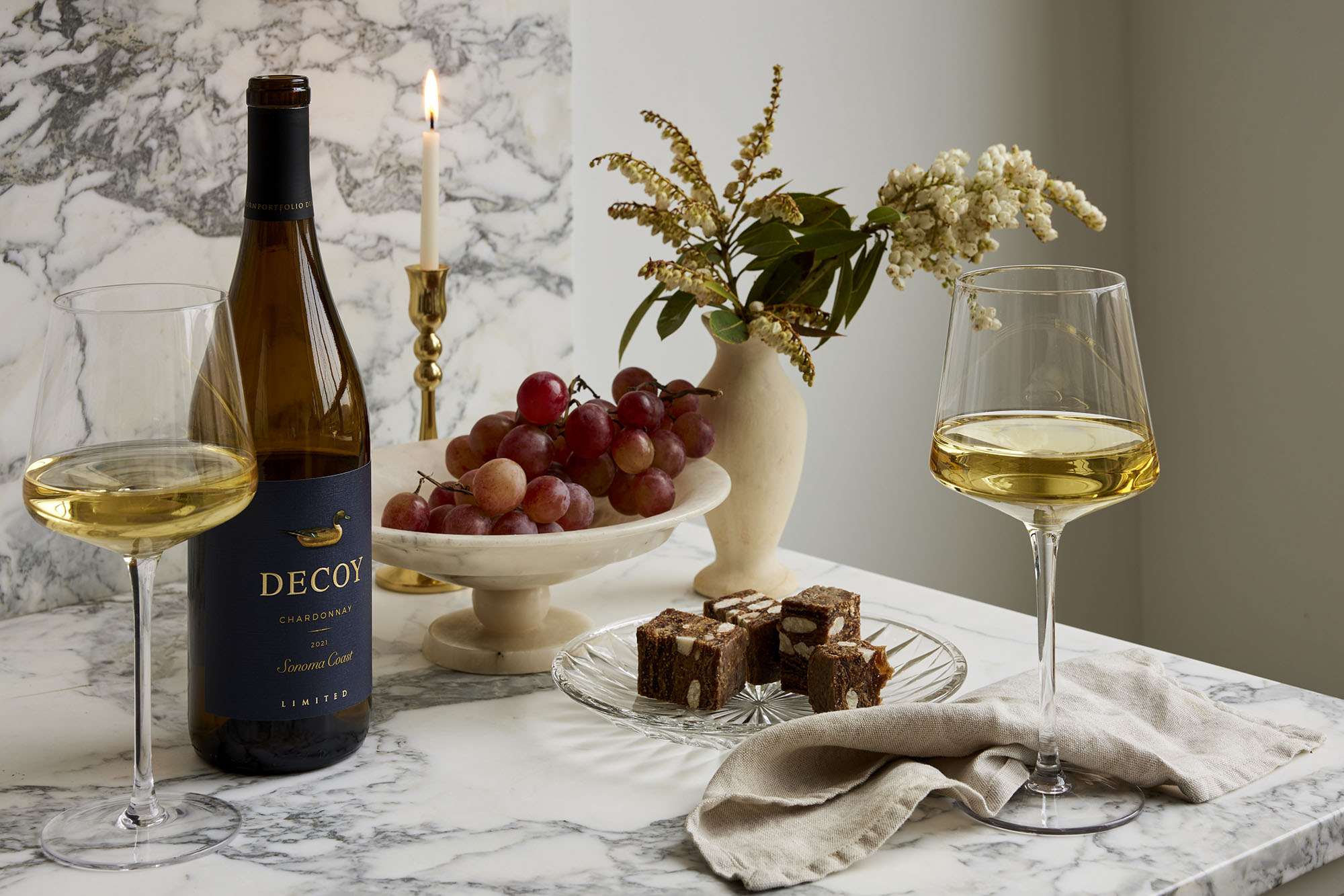 A sophisticated scene featuring a marble table adorned with a bottle of white wine and a glass of wine. The table is set with delicate floral arrangements and glass dishes filled with figs and snacks, creating an elegant ambiance enhanced by candlelight.