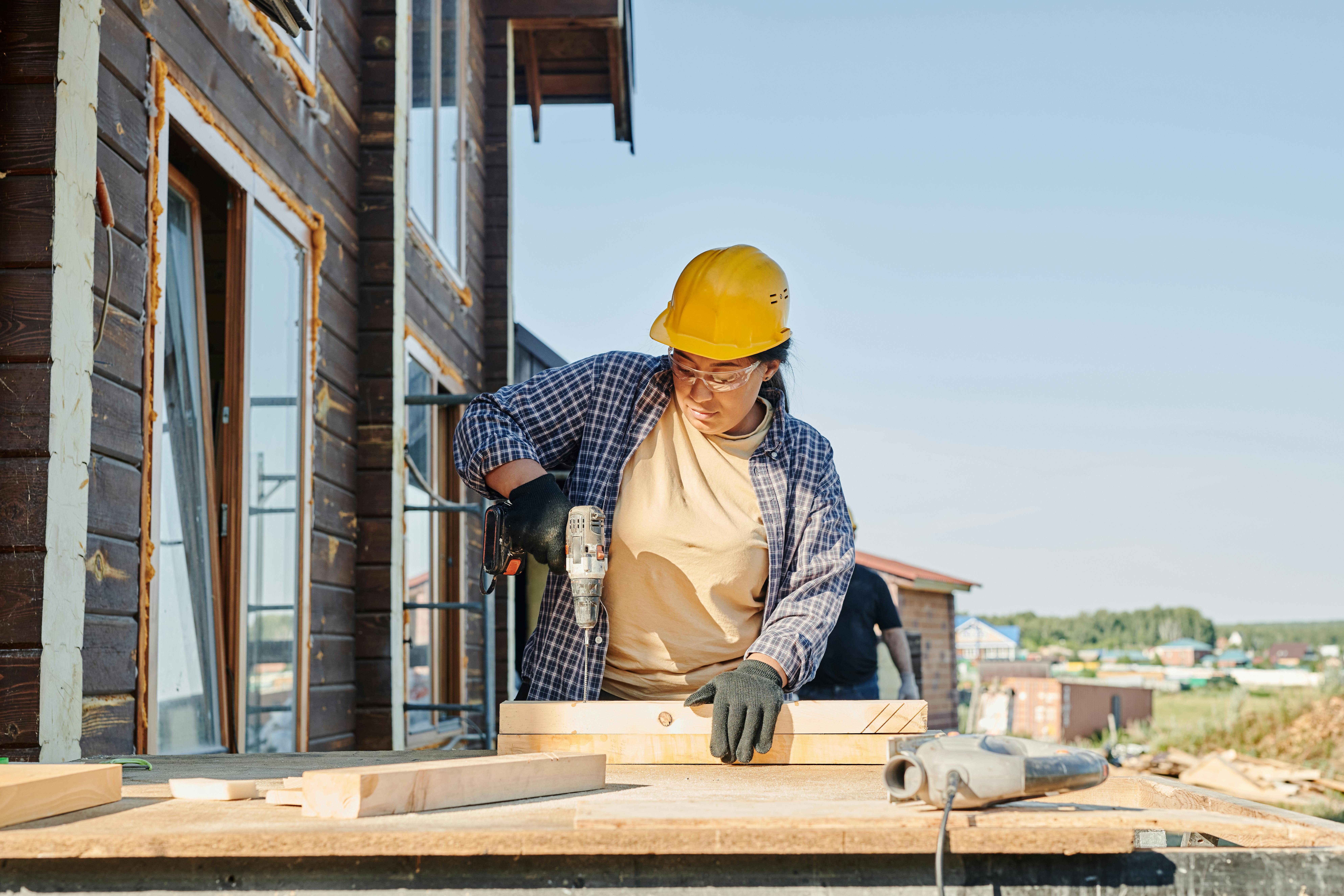 A construction worker in a yellow hard hat and safety goggles uses a drill on wood outside a wooden house under a clear blue sky.
