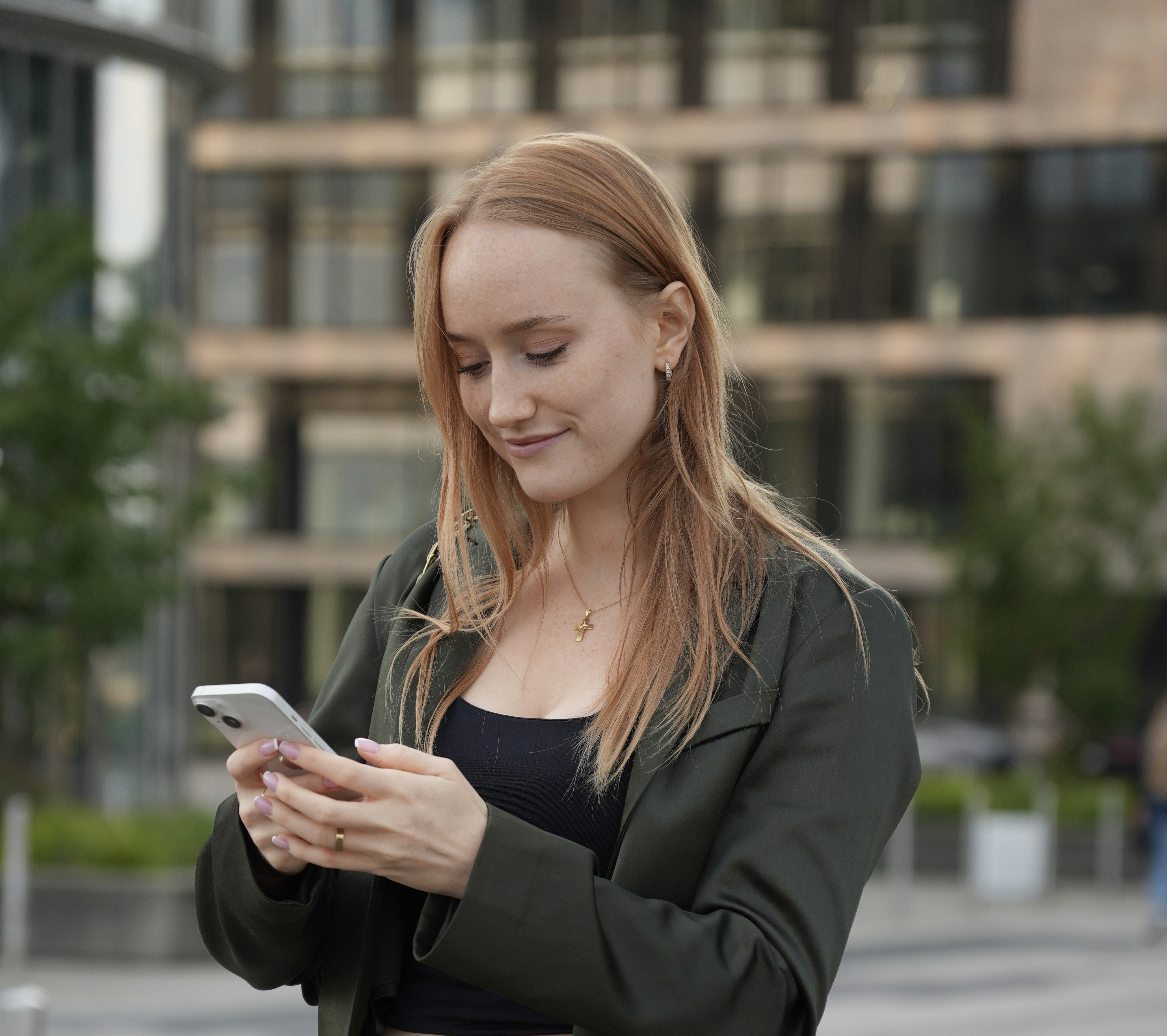 Two smiling people outdoors, one in a pink jacket holding a white phone, the other in blue with a buzz cut holding a blue phone, under a clear sky.