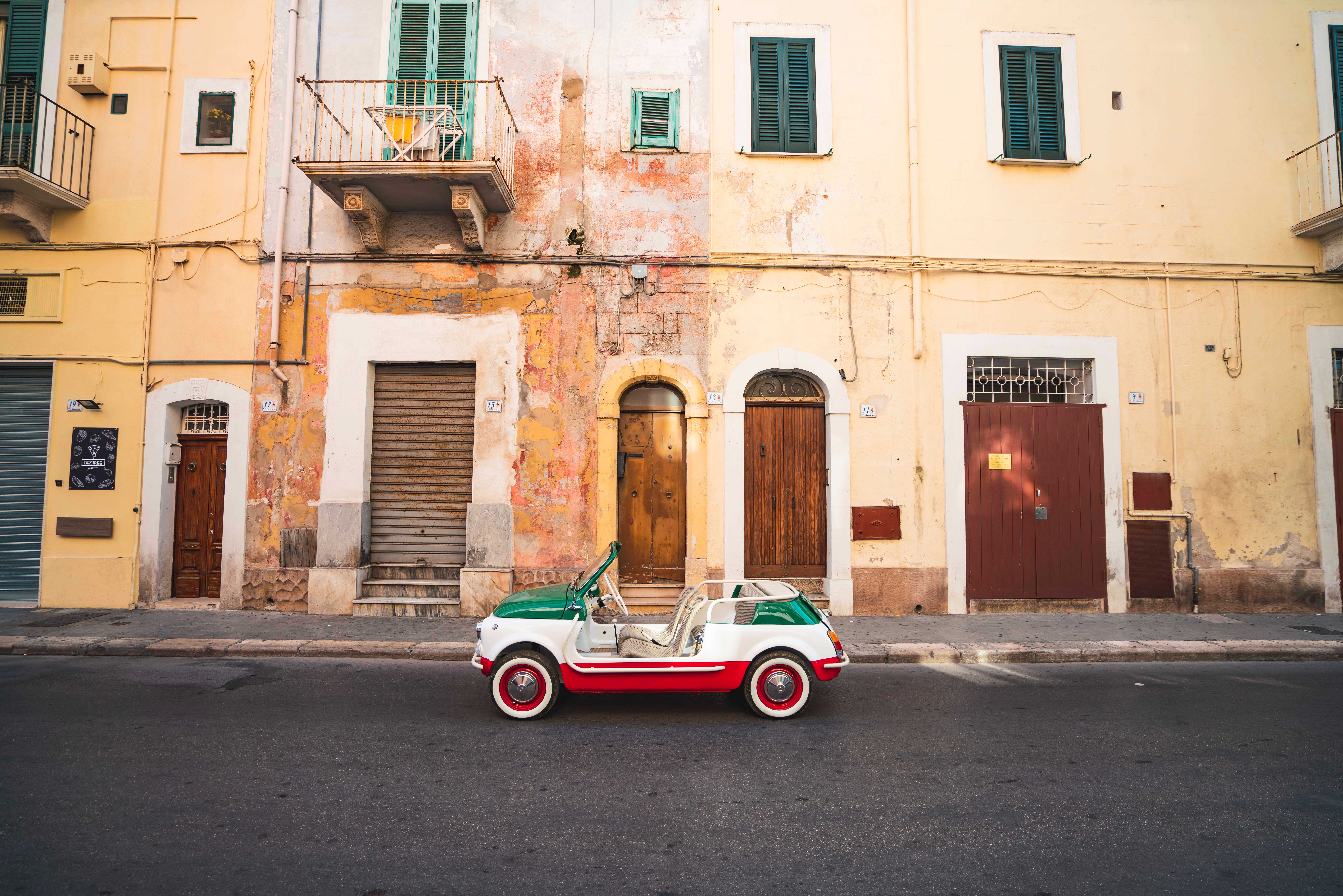 A small, vintage car painted in the colors of the Italian flag—green, white, and red—is parked on a street in front of a row of old, colorful buildings with various wooden doors.