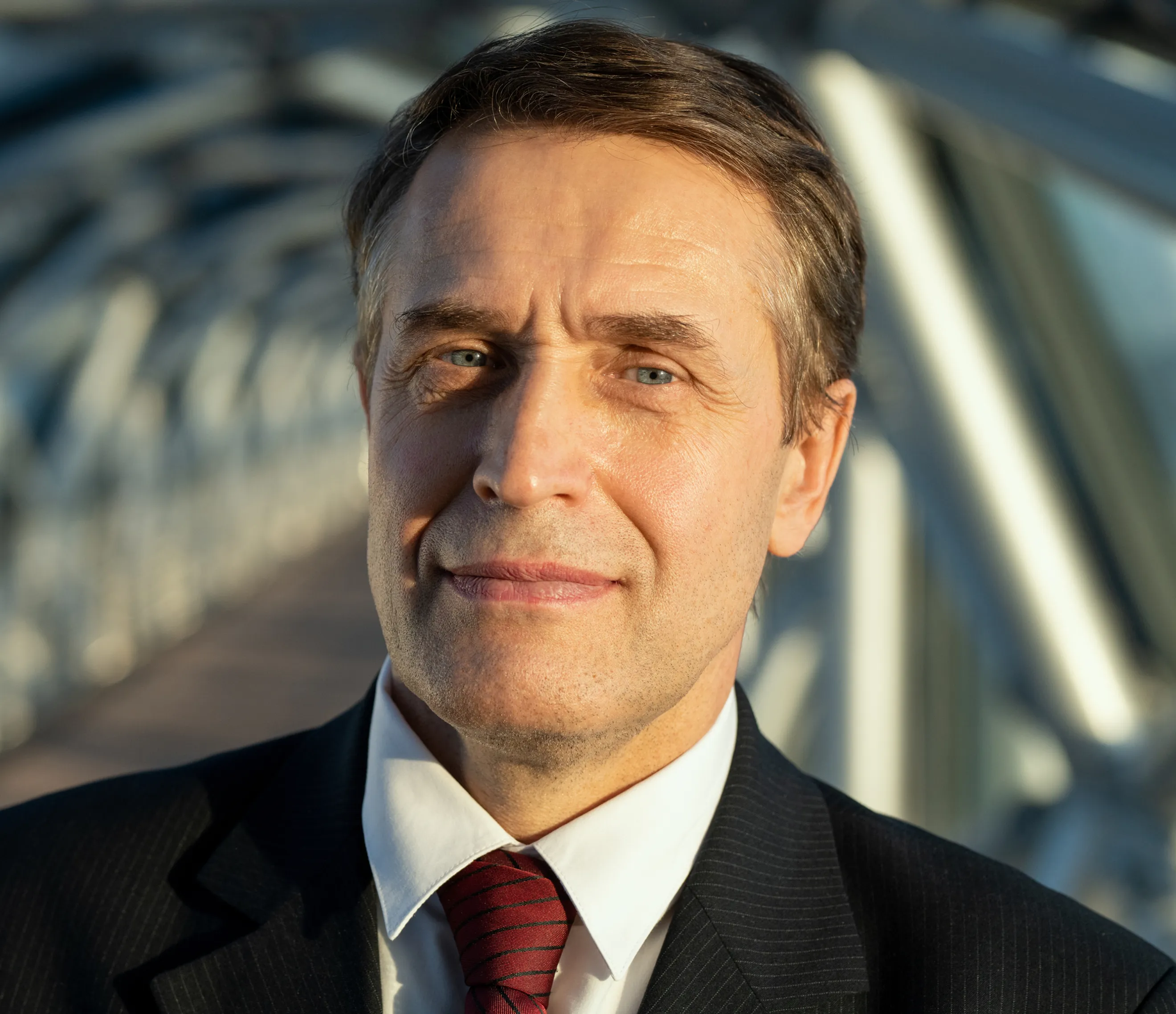 Close-up portrait of a confident male executive in a dark suit and maroon tie with a blurred architectural background