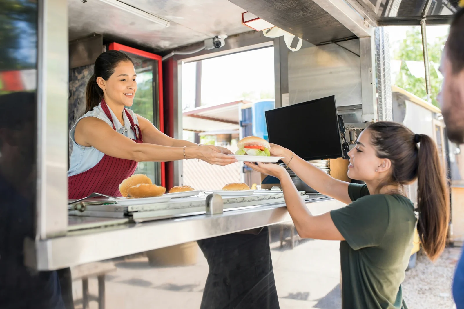 Food truck worker in red apron hands burger to smiling customer at service window.