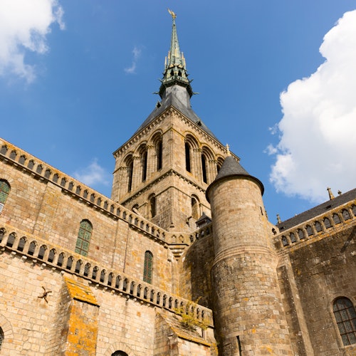 Stone building with tall, ornate tower, arched windows, and turrets under a bright blue sky with scattered white clouds.