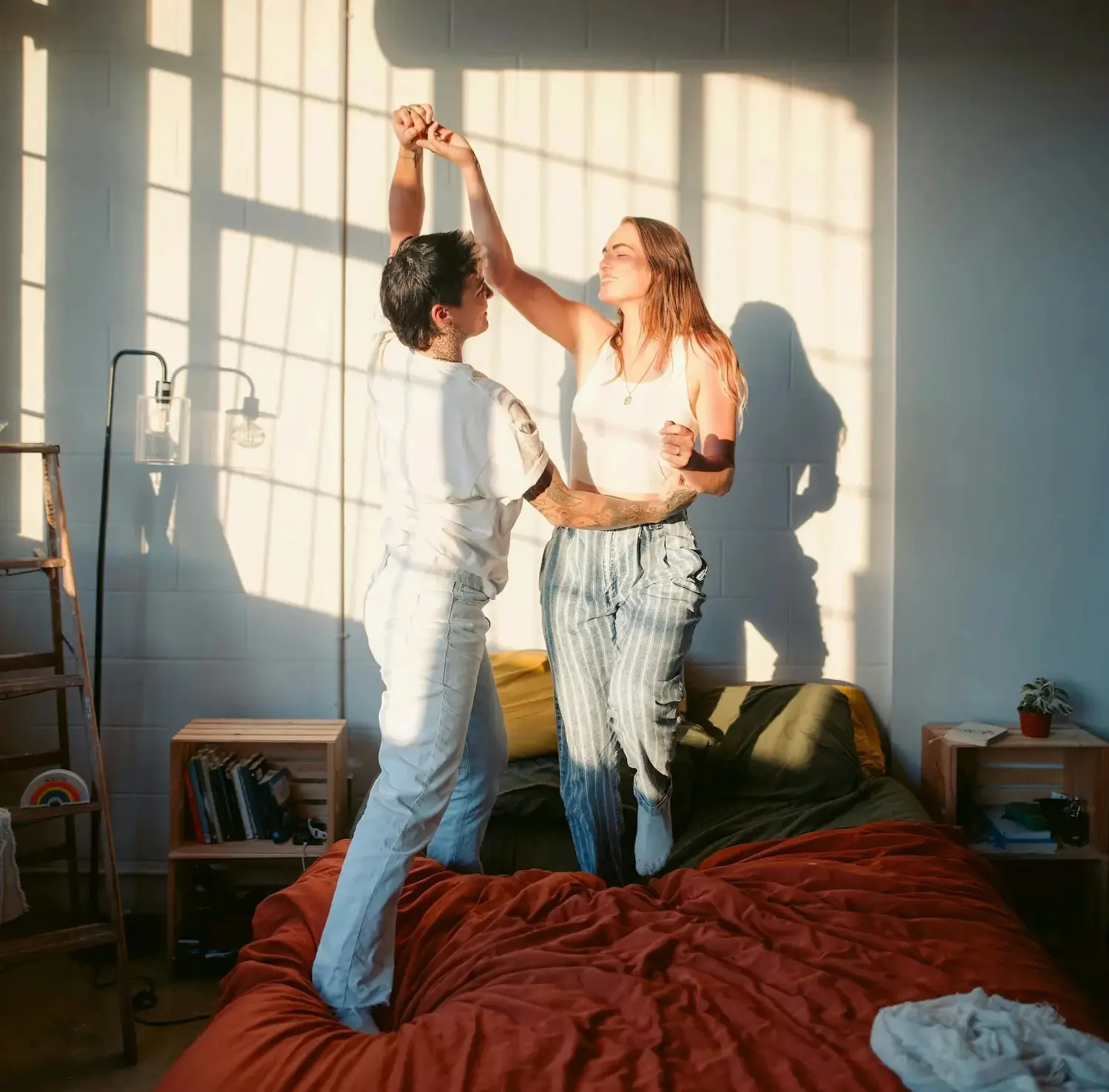 Two queer women dancing together on a bed in a sunlit bedroom, embracing joy and intimacy in a healthy same-sex relationship.