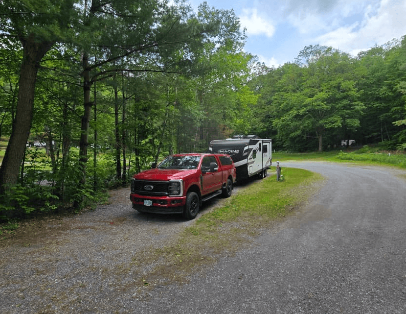 Red pickup truck towing a white camper on a gravel road through a lush green forest