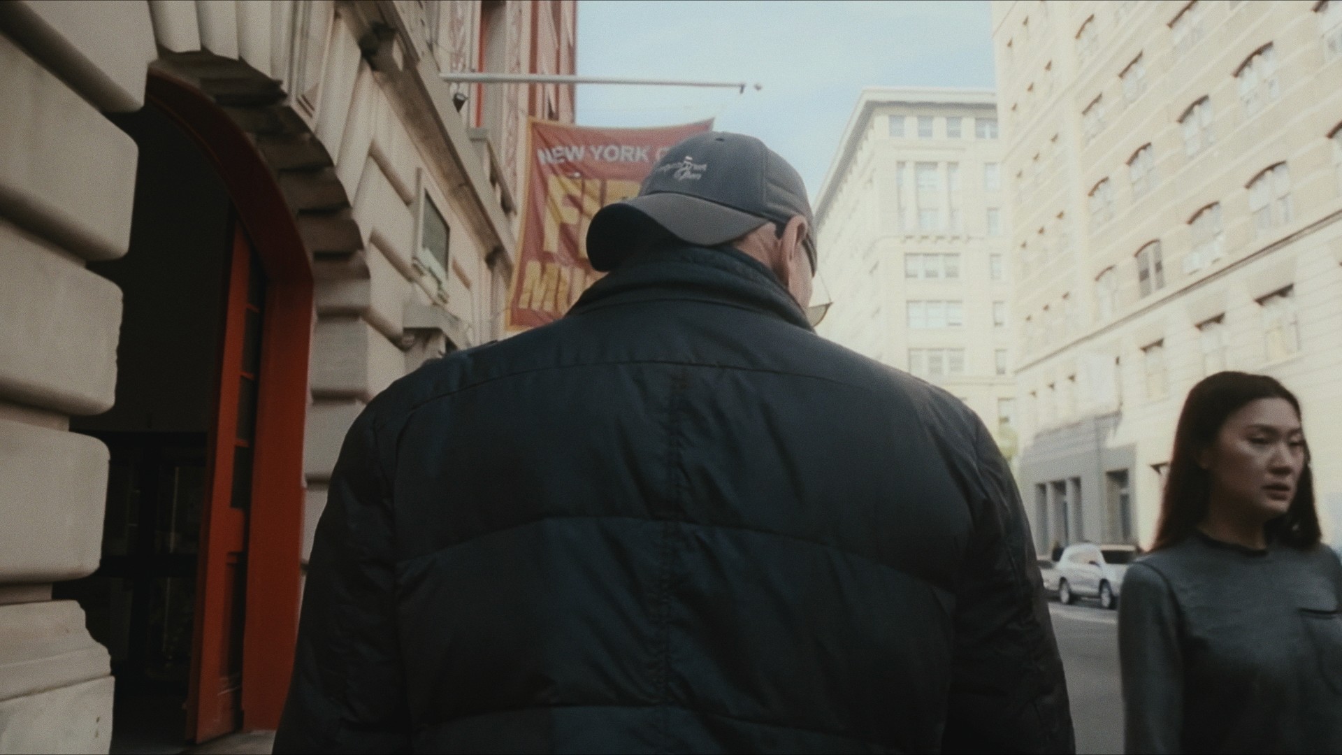 A man in a cap and dark jacket walking a city street, seen from behind, a woman beside him, Maddman