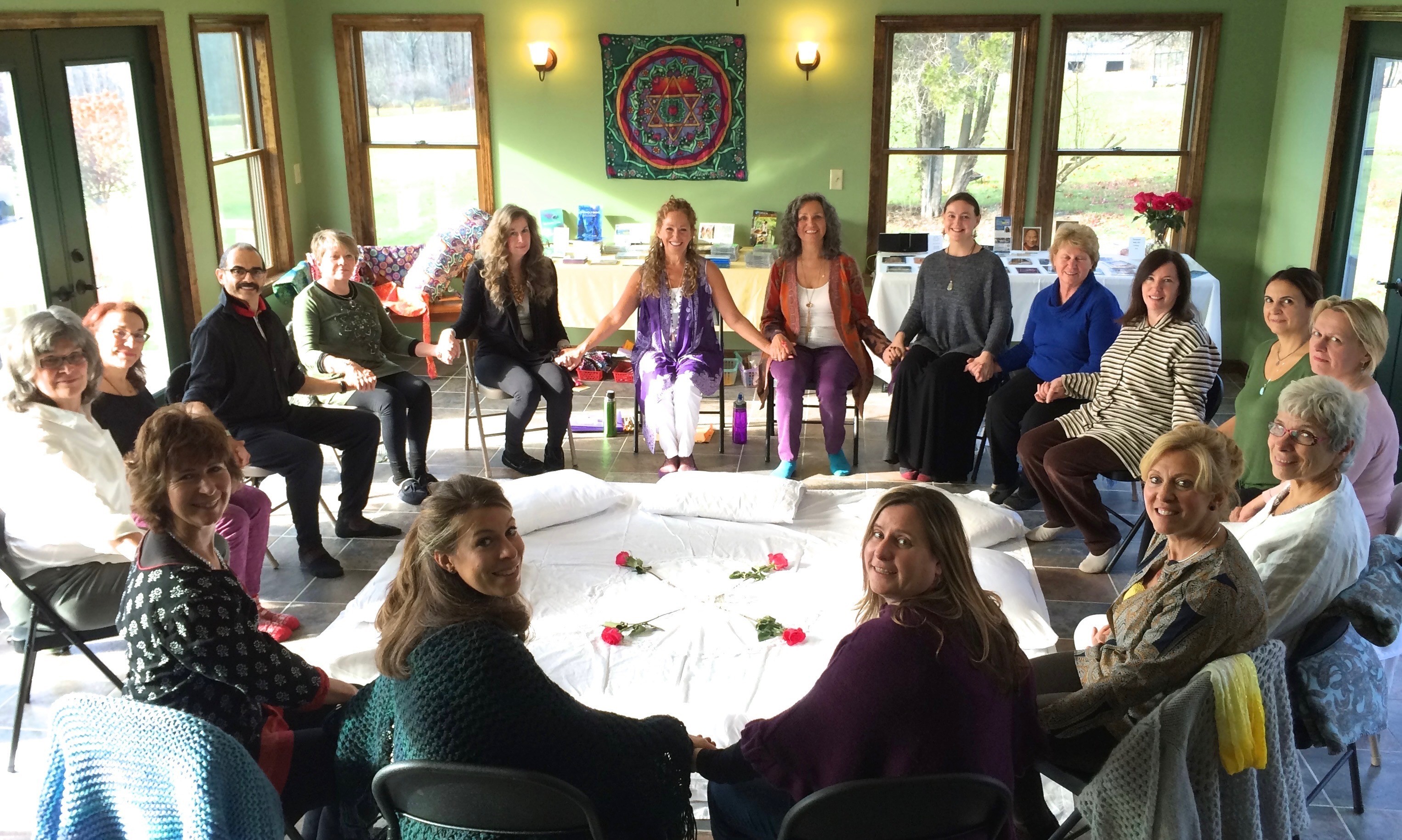 A smiling man with curly hair is sitting at a desk, looking at the camera, with a laptop and a lamp in the background.