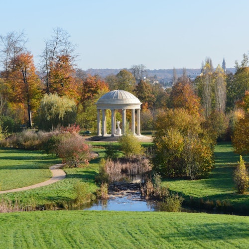 Un parque pintoresco con un quiosco abovedado rodeado de árboles con follaje otoñal, un sendero serpenteante y un pequeño arroyo.
