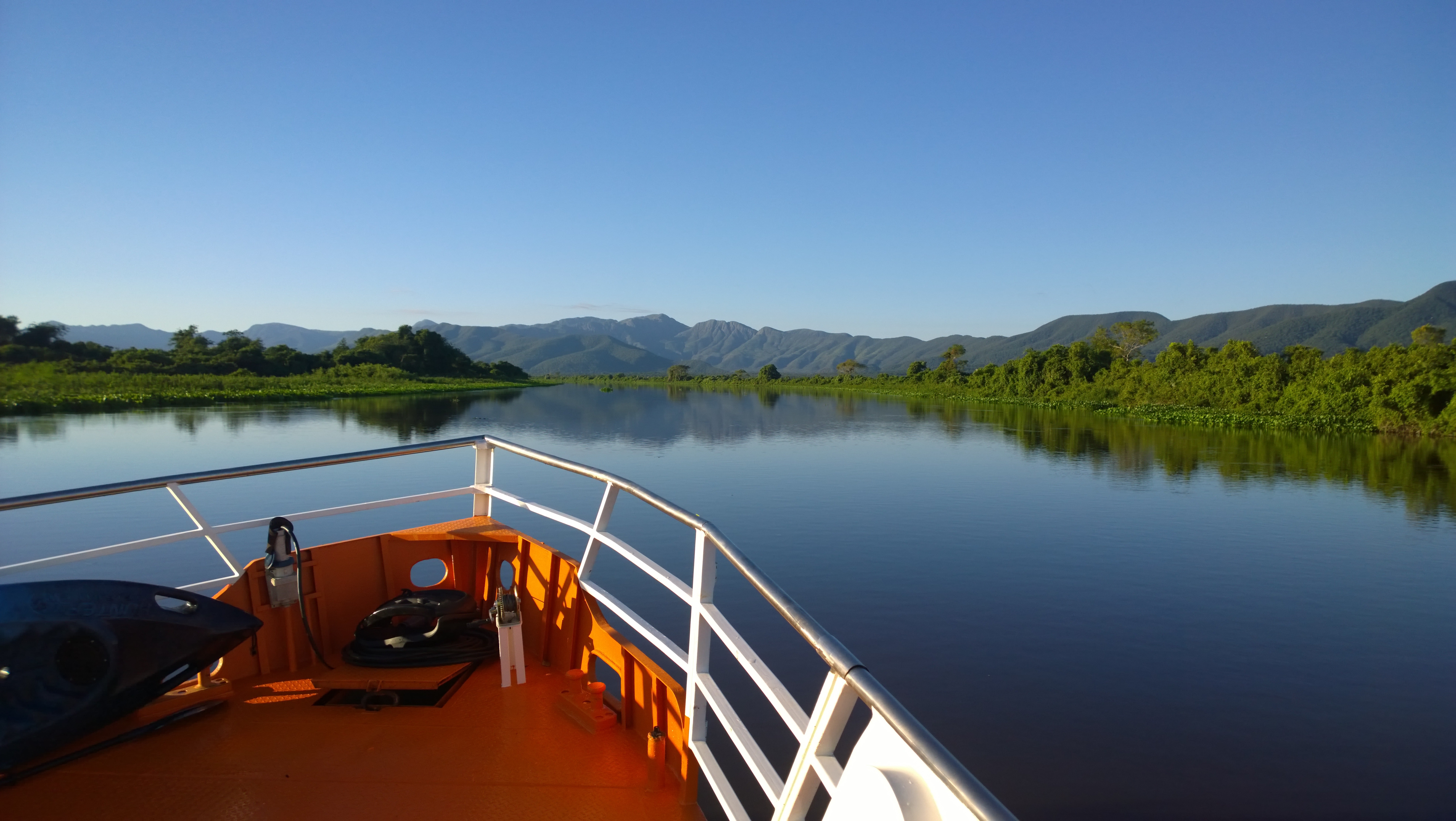 Blick vom Bug eines Expeditionsschiffs auf dem Rio Paraguai im West-Pantanal – die grüne Uferlandschaft und die majestätische Serra do Amolar spiegeln sich im ruhigen Wasser unter strahlend blauem Himmel.