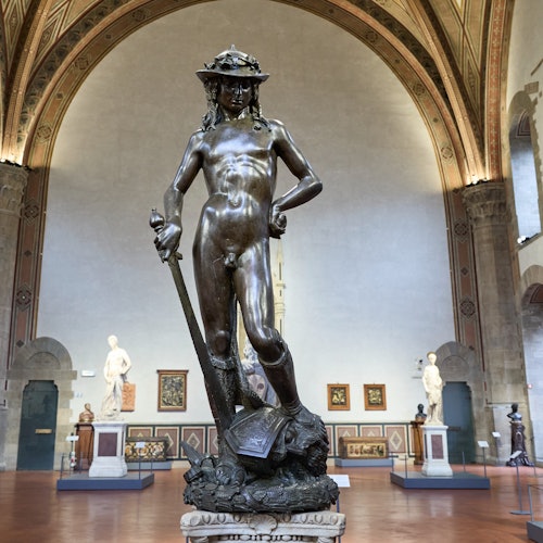 Une statue en bronze de David, réalisée par Donatello, se dresse dans une salle de musée aux hauts plafonds voûtés, entourée d'œuvres d’art.