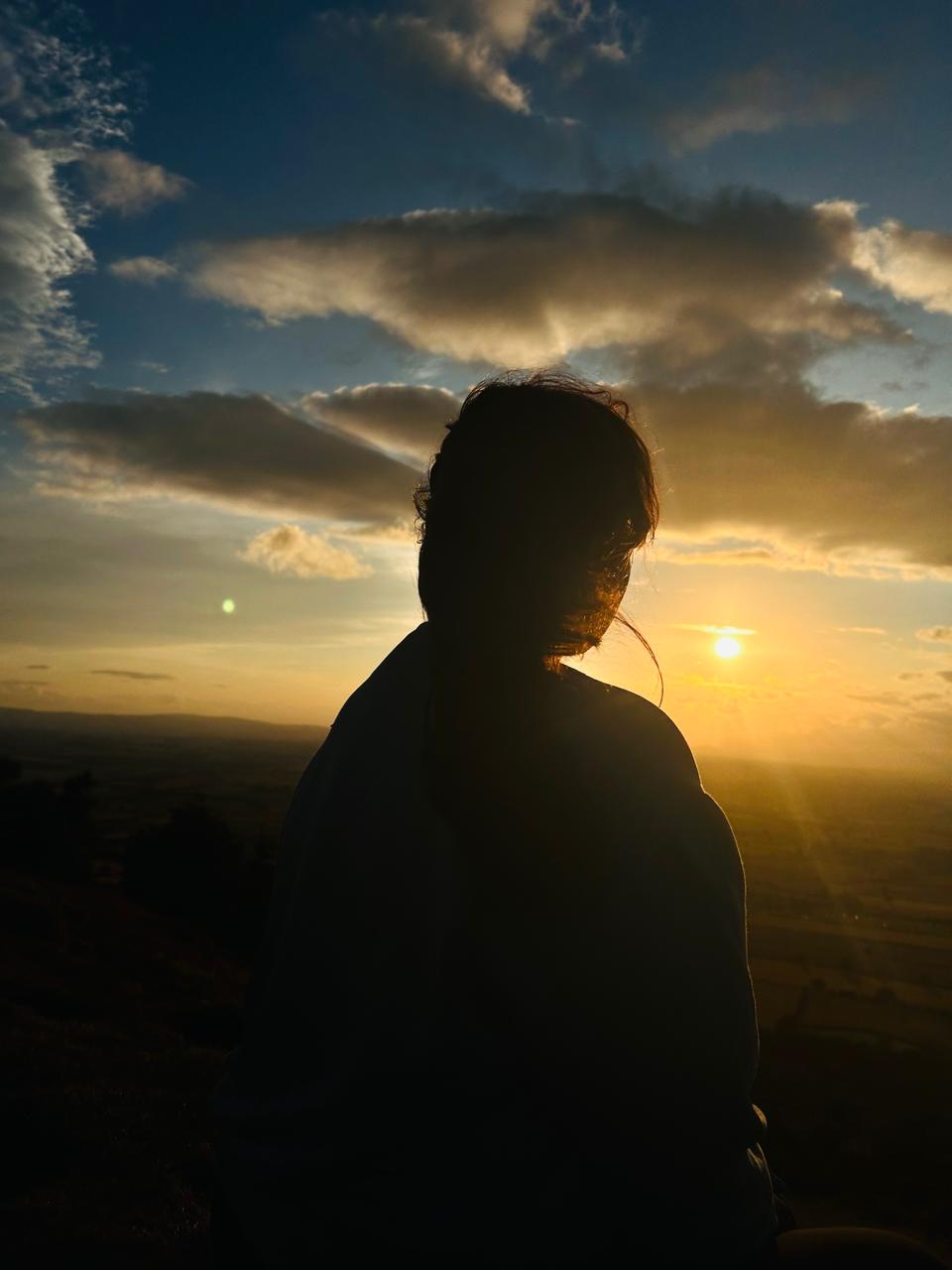 Una persona con una camiseta negra y pantalones de color claro está de pie en la cima rocosa de una montaña al atardecer, sosteniendo una cámara frente a su cara para tomar una foto. Un mar de nubes se extiende detrás de ellos, iluminado por la luz dorada del sol poniente.