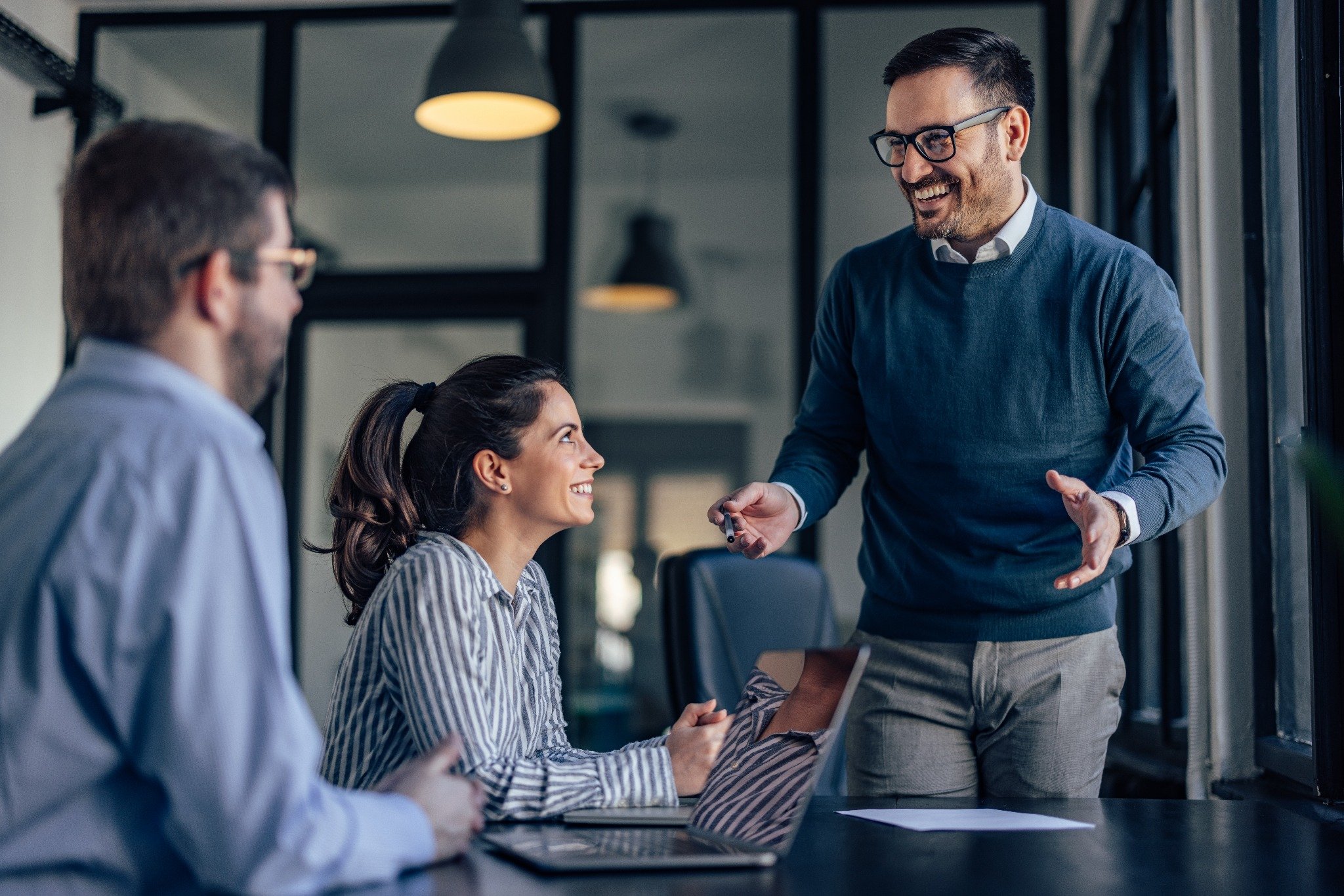 Zwei Büromitarbeitende sitzen an einem Holztisch, auf dem ein Laptop und viele Dokumente liegen. Die beiden Kolleg:innen geben sich einen High Five und lachen sich an.