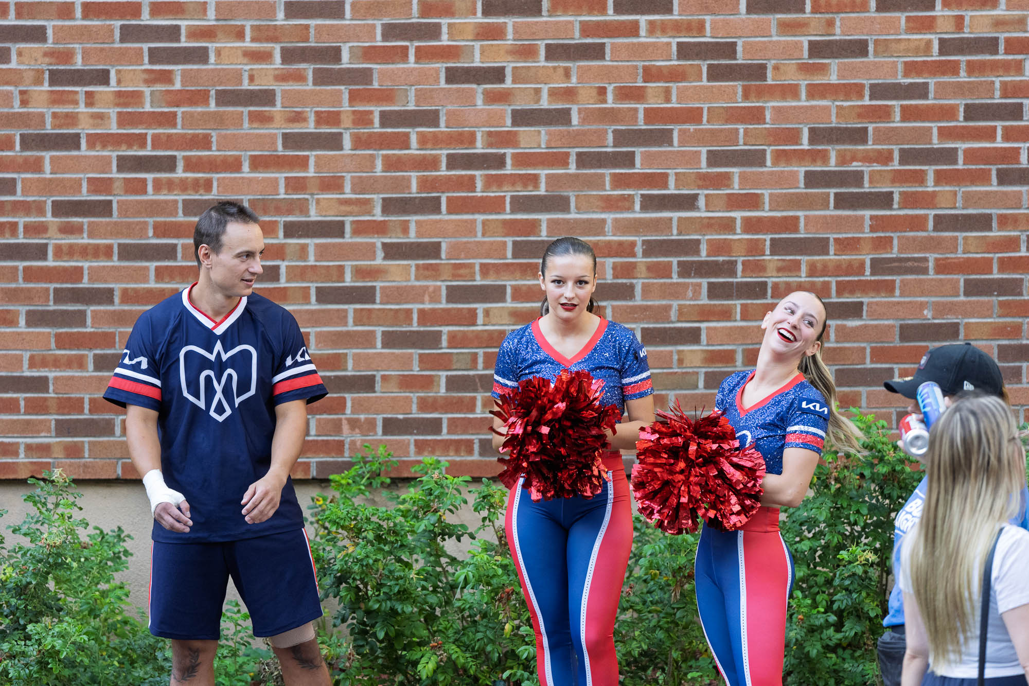 05_septembre_2026Quebec_20266779.jpg — Un joueur des Alouettes en maillot d’entraînement bleu marine discute avec deux cheerleaders aux pompons rouges devant un mur de briques, à l’extérieur du stade Percival-Molson.