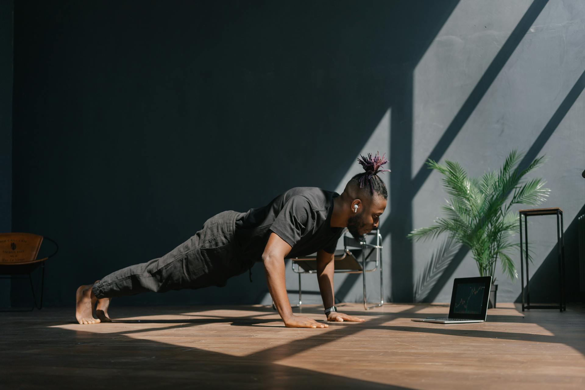 A man with dark clothes performs a push-up at home on a wooden floor next to a laptop.