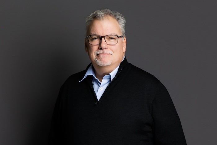 A man with glasses and a brown shirt, crossing his arms.