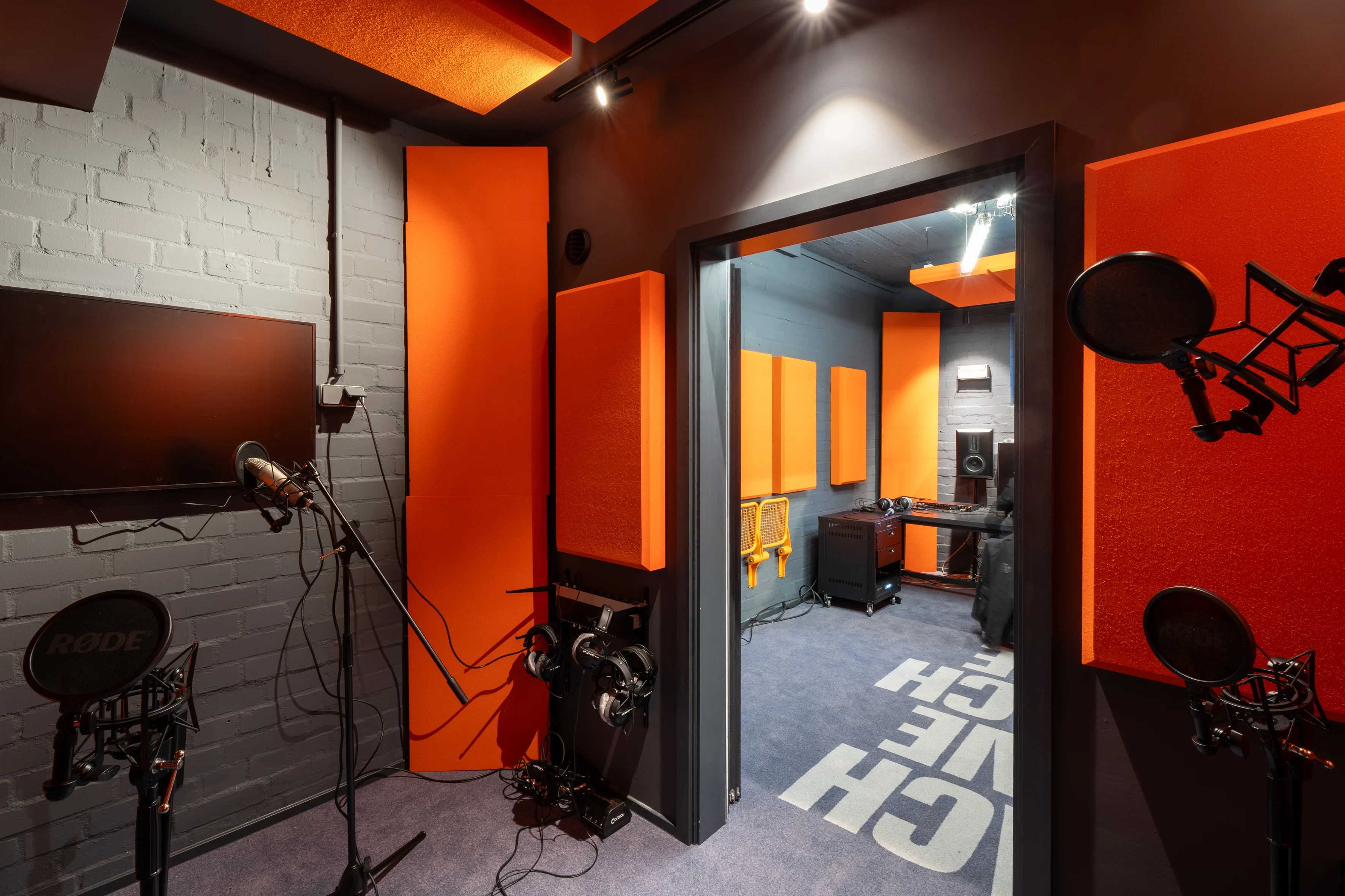 View from the vocal booth at SNIPES BASEMENT with microphones and headphones into the orange-paneled control room with studio monitors and mixing desk.