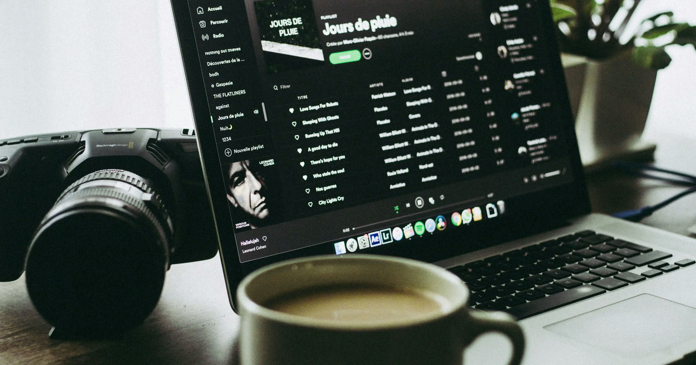 Laptop playing a Spotify playlist, with a coffee cup and a professional camera on the desk.