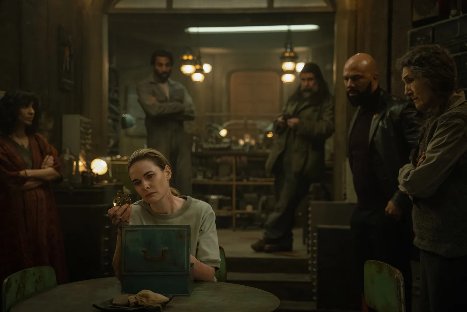 In a dimly lit, vintage room filled with rustic décor, a thoughtful woman sits at a round table inspecting an object, surrounded by five people who appear engaged in a serious discussion, under the glow of warm overhead lamps.