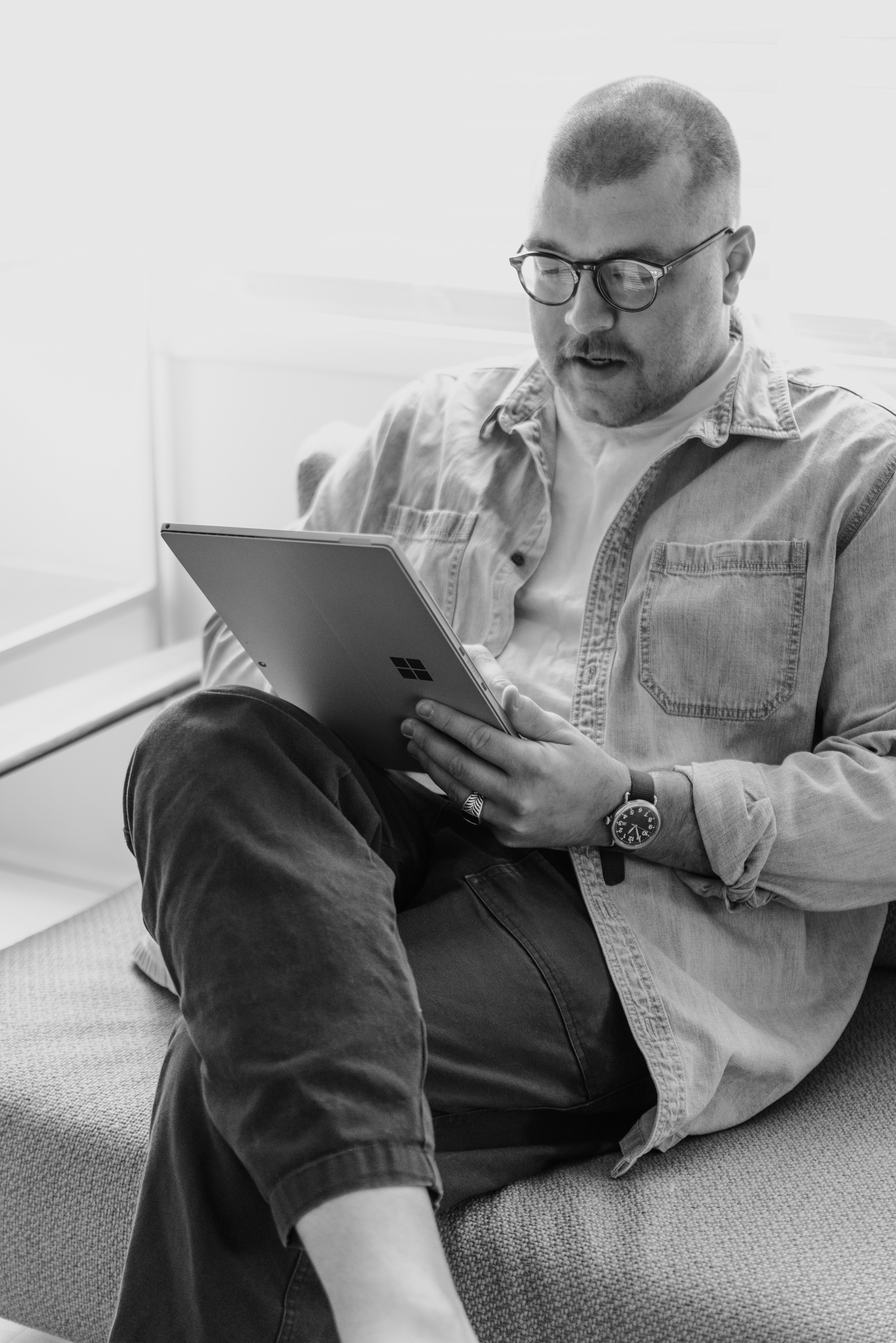 A man wearing glasses sits on a couch, focused on using a tablet in a black and white setting.