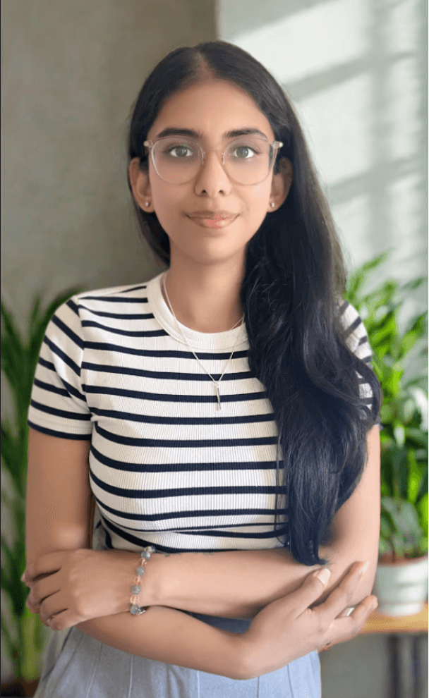 Vrinda in black and white striped top with hands folded near waist, standing against a backdrop of plants, introducing her therapeutic approach.