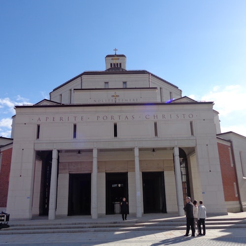 Фасад церкви с колоннами и крестом наверху. Четыре человека стоят перед ним под ясным голубым небом.