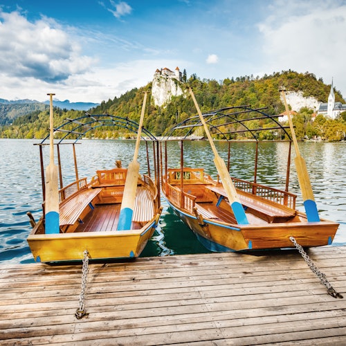 שתי סירות עץ עם משוטים עוגנות באגם, עם נוף גבעתי ויער ברקע וטירה ברקע.
